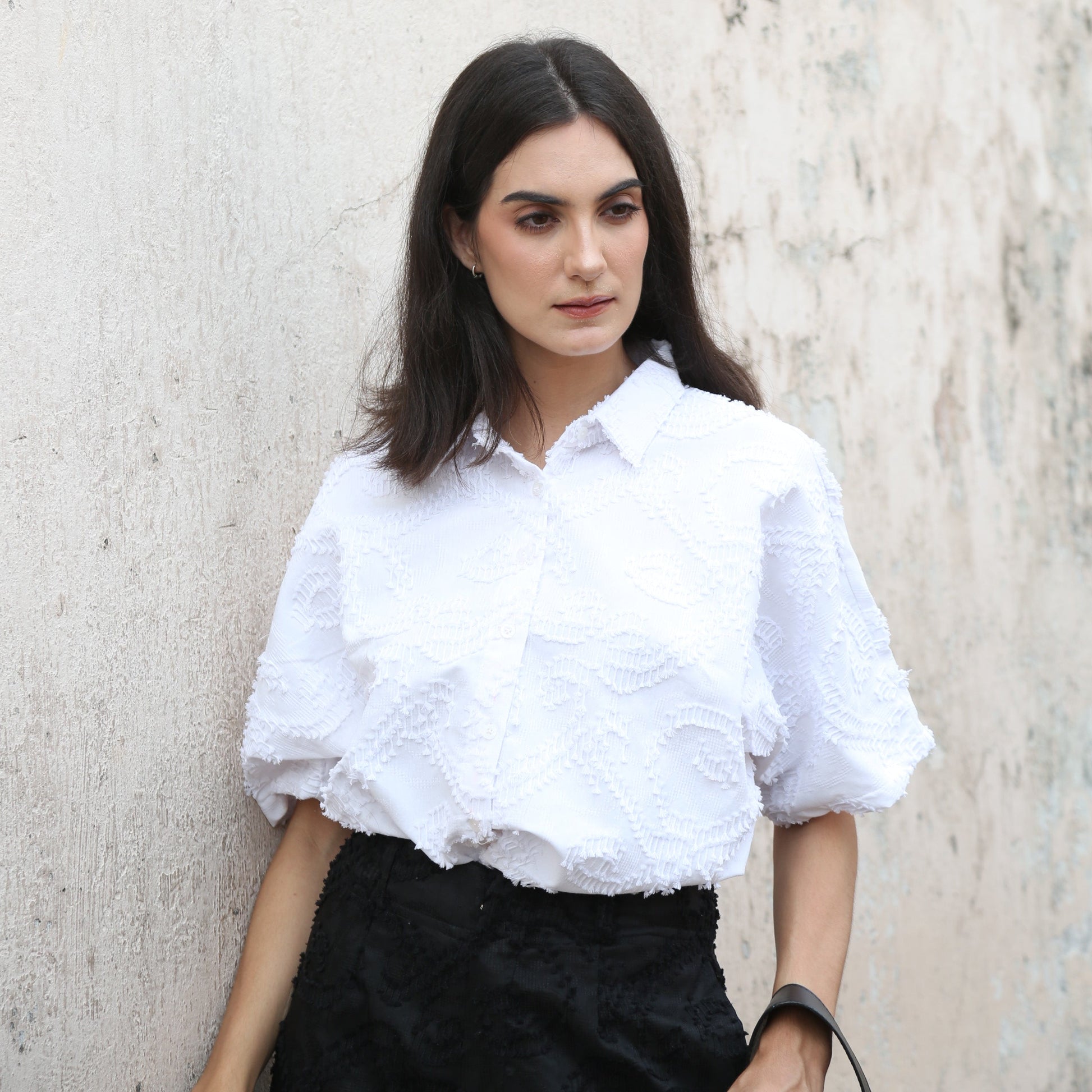 A woman standing against a textured wall wearing a white shirt with short sleeves and black pants.