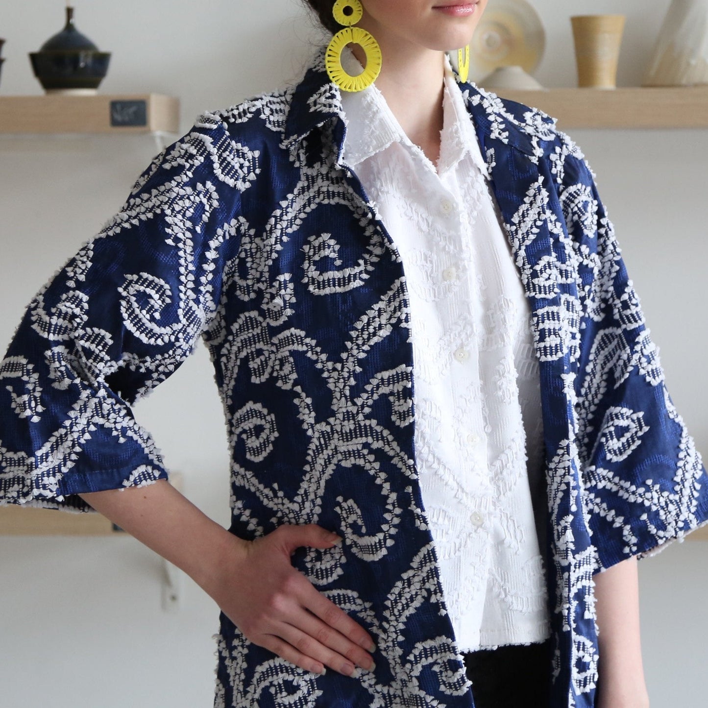 A woman standing in a studio setting, wearing a blue and white geometric patterned long outerwear with a collared neck and two pockets. She is also wearing a white shirt and yellow earrings.