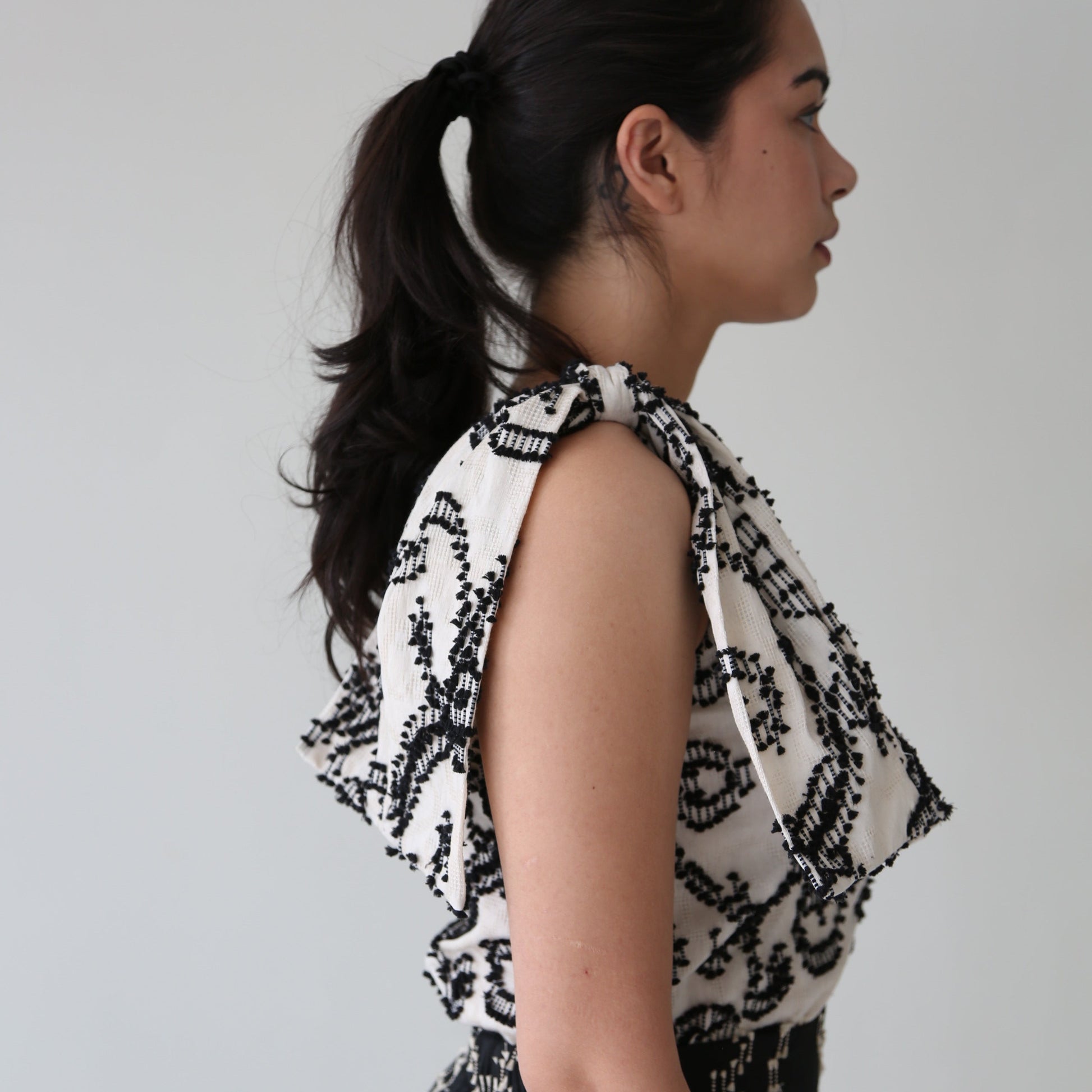 A woman wearing a sleeveless top with a dramatic bow on one shoulder and a round neckline, displayed in a side profile view.