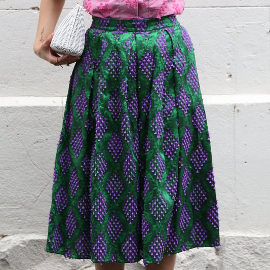 A person wearing a knee-length skirt with a geometric pattern in green and purple, paired with white shoes and teal socks.