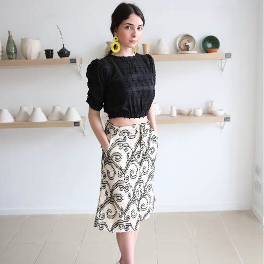 A woman standing in a studio setting, wearing an A-line below the knee skirt with front slit, in a floral pattern, paired with a black top and white shoes. The background displays a variety of ceramic art pieces.