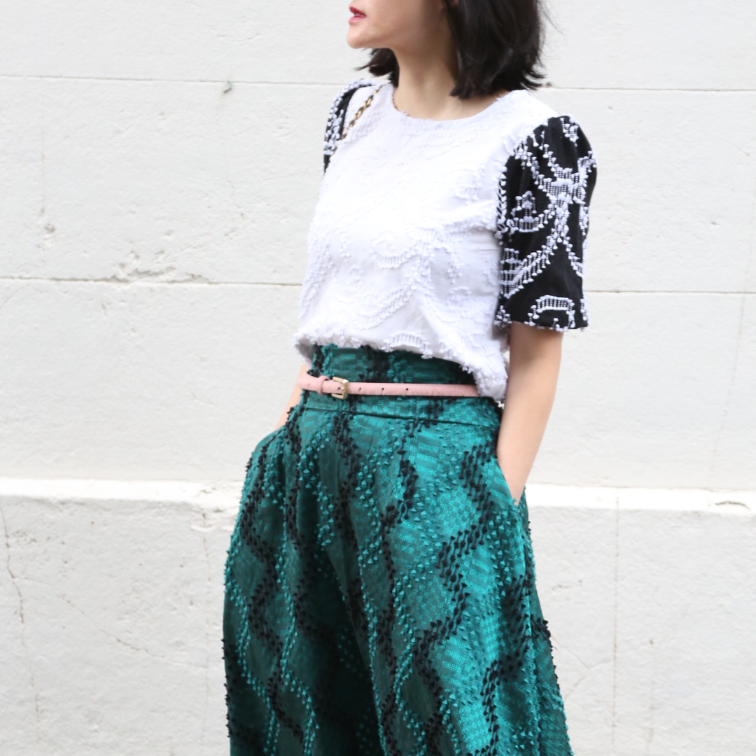 Person wearing a white top with black patterns and a green patterned skirt against a white wall.