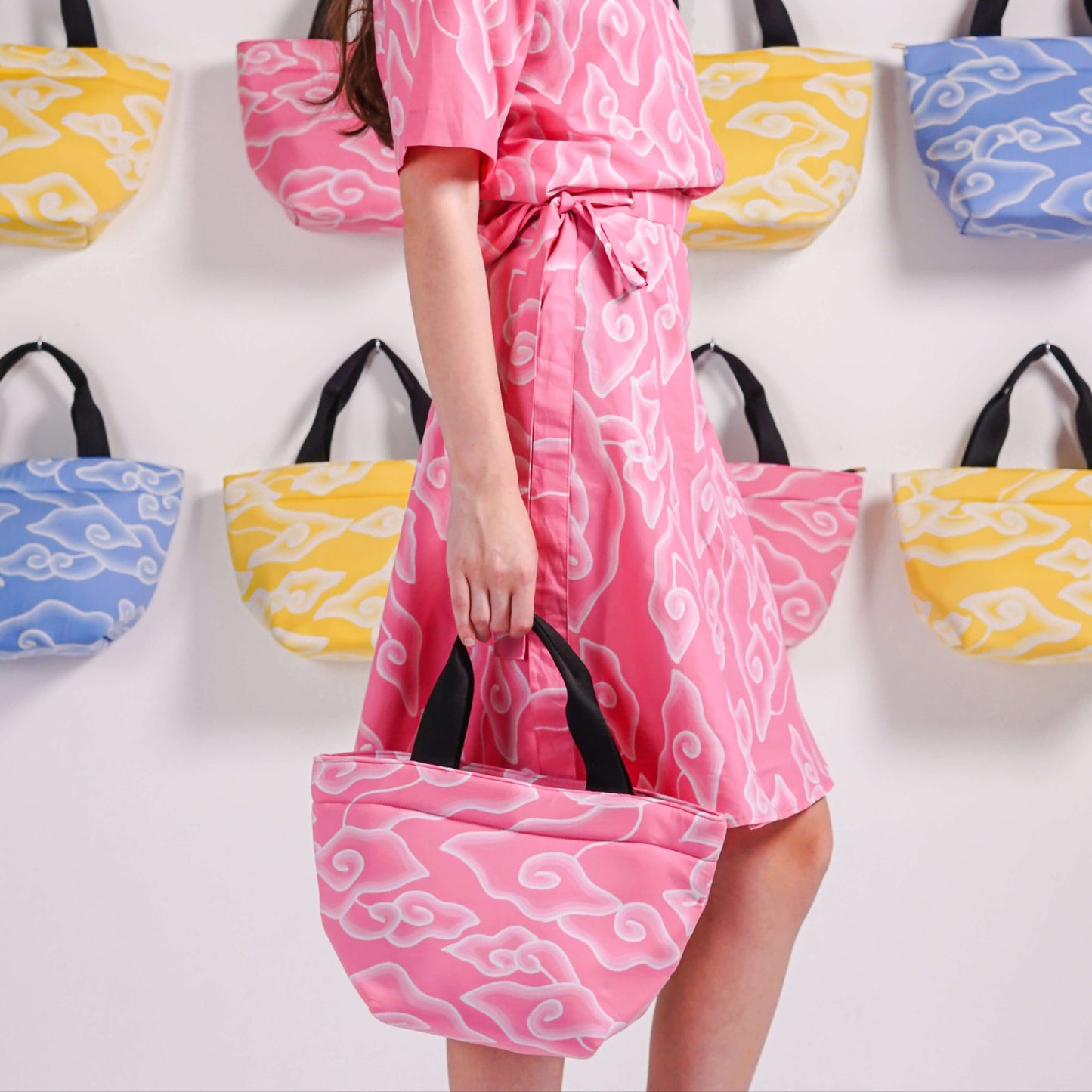 A series of tote bags with colorful hand-painted batik patterns displayed on a wall, with a woman standing in front of them carrying a bag with a similar pattern.